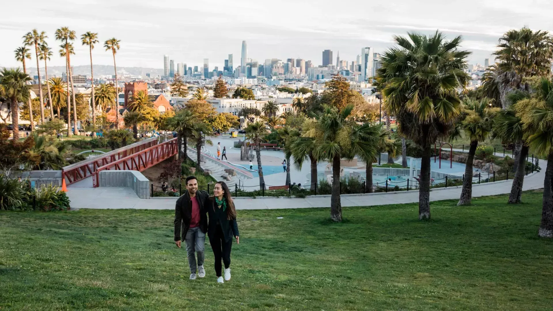 一对夫妇走向镜头，Dolores Park和贝博体彩app的天际线在他们身后.